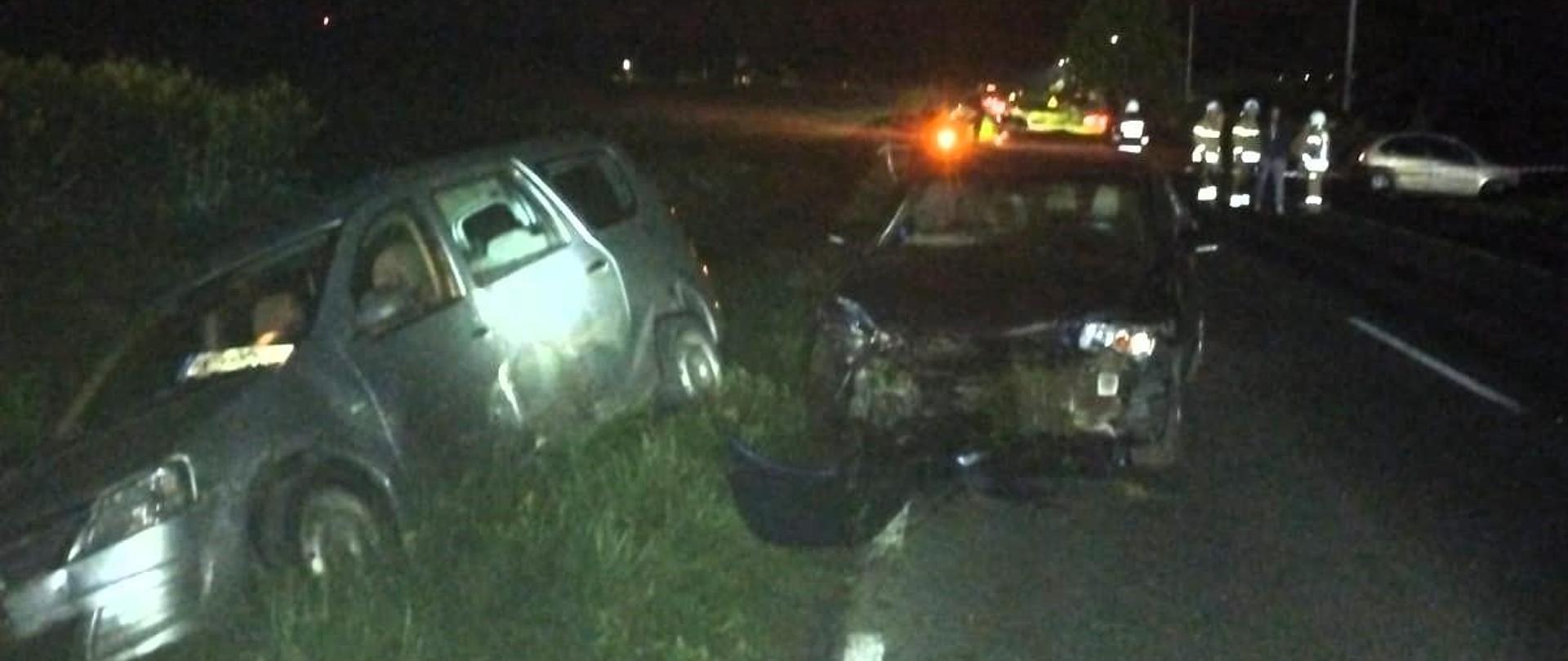 Zdjęcie przedstawia drogę wojewódzką: na pierwszym planie dwa spośród czterech uszkodzonych samochodów, w głębi samochody służb ratowniczych i strażacy. 