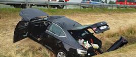 samochód czarny po dachowaniu w rowie w tle samochody strażackie oraz policyjny radiowóz