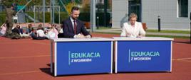 Na boisku szkolnym stoi dwa stoły na których widnieje napis Edukacja z wojskiem. Za stołem siedzi minister MON i Minister edukacji. Podpisują dokumenty. W tle widać dzieci siedzące na boisku. 