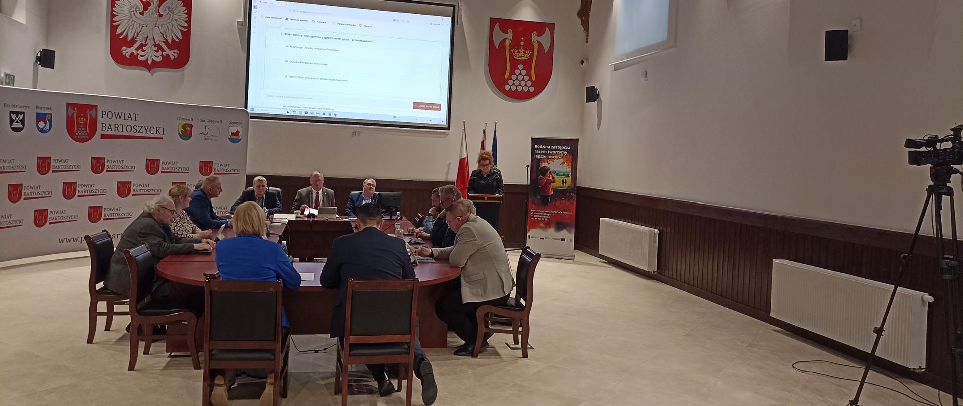W Sali obrad znajduje się duży owalny stół, przy którym siedzi kilkanaście osób biorących udział w sesji Rady Powiatu. Na ścianie za nimi widoczny jest duży ekran z projekcją dokumentu. Po bokach wiszą herb oraz godło Polski. Z prawej strony stoi mówiąca osoba przy mównicy. W tle widoczne są flagi