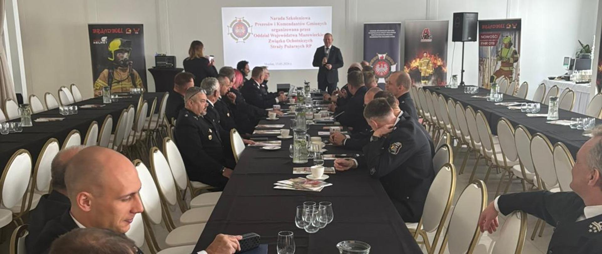 Sala konferencyjna podczas narady szkoleniowej prezesów i komendantów gminnych OSP. Uczestnicy w mundurach siedzą przy długich stołach, a na końcu sali trwa prezentacja. 
