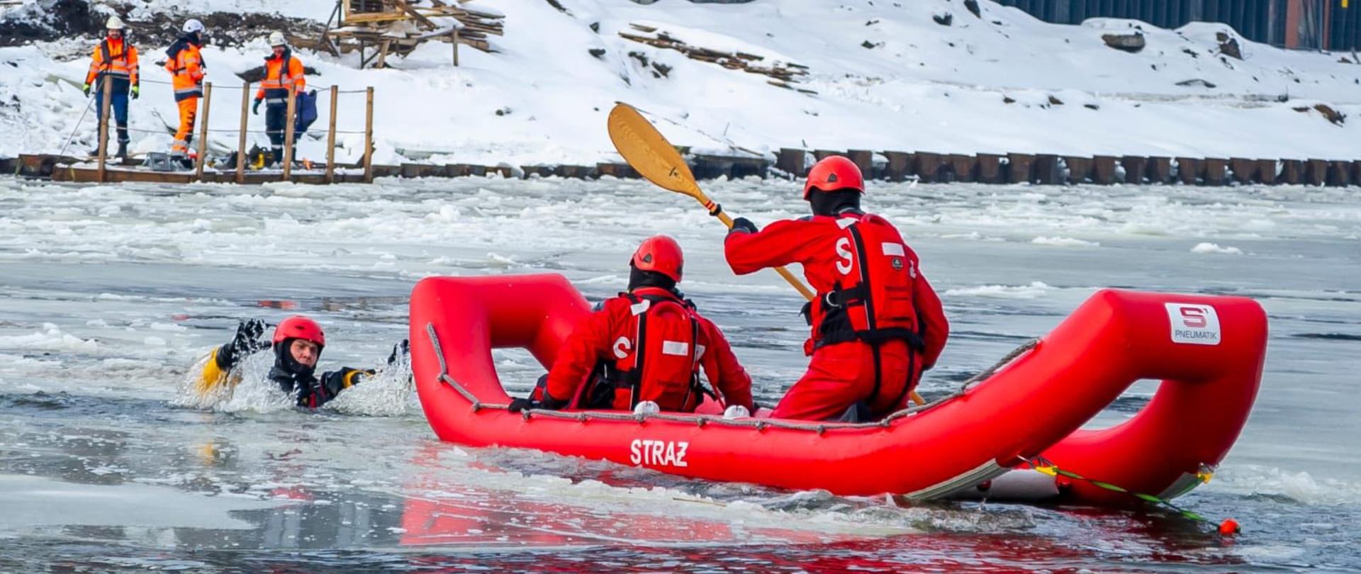 Na zamarzniętym akwenie trwa akcja ratownicza. Dwóch ratowników w czerwonych kombinezonach i kaskach siedzi w dmuchanej łodzi i podpływa do osoby, która wpadła do lodowatej wody. Wokół niej widać pokruszony lód. Na ośnieżonym brzegu stoi kilku ratowników w pomarańczowych strojach, obserwujących i koordynujących działania. Scena odbywa się w zimowych, surowych warunkach i pokazuje profesjonalną pracę służb ratowniczych.