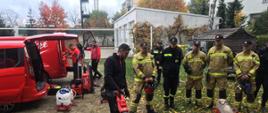 Grupa strażakow stoi w rzędzie, przed mężczyzną prezentujęcym sprzet techniczny. W tle widoczne czerwone samochody, z których dwóch mężdzyzn wyciąga kolejny sprzęt do prezentacji