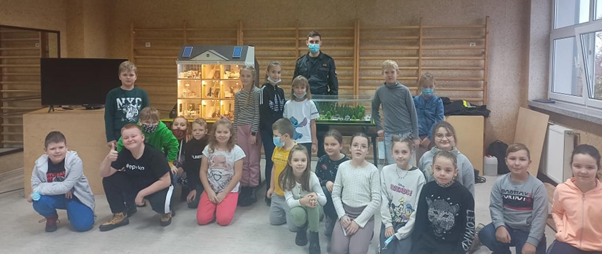 Zdjęcie grupowe dzieci i strażaka, w tle symulator i makieta. Wszyscy znajdują się w sali.