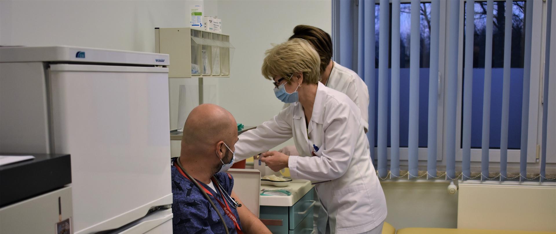 Pomieszczenie, w którym wykonywane jest szczepienie. W fotelu siedzi szczepiona osoba, obok personel medyczny przygotowuje dawkę szczepionki.