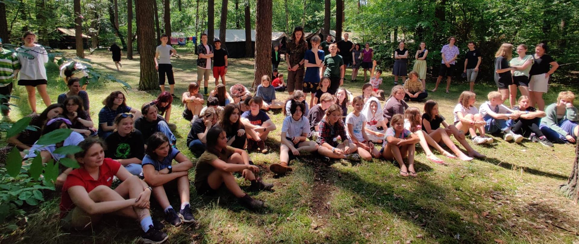 zdjęcie przedstawia siedzącą grupę dzieci podczas zajęć na wolnym powietrzu 