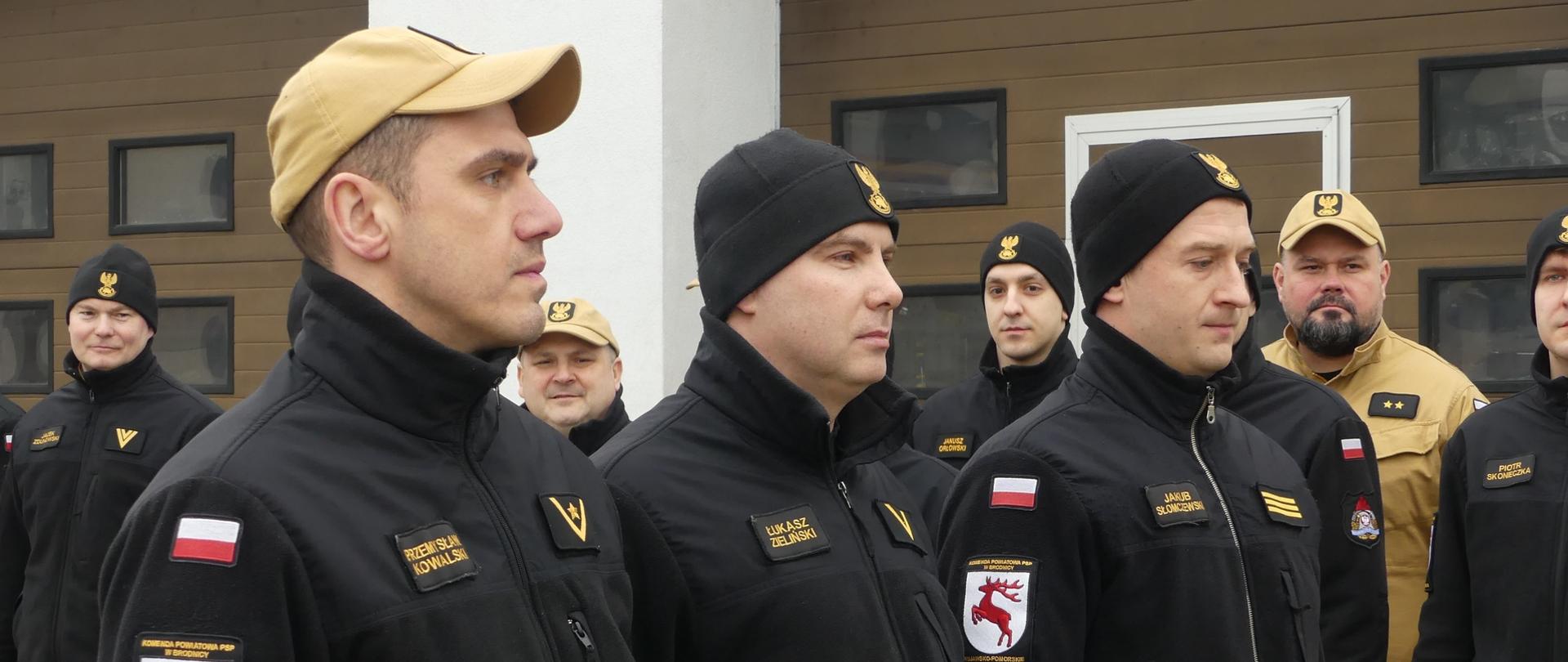 Zdjęcie przedstawia trzech funkcjonariuszy PSP w Brodnicy mianowanych na wyższe stanowiska służbowe. Ubrani w stroje służbowe oczekują na wręczenie decyzji przez Komendanta Powiatowego. Za nimi w dwuszeregu stoją inni funkcjonariusze Brodnickiej Komendy.