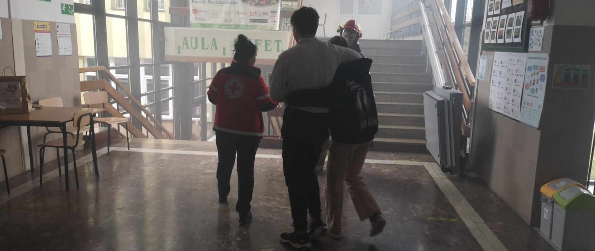 Na zdjęciu widać kobietę w bluzie PCK oraz dwójkę uczniów którzy wyprowadzają się pod ramię ze szkoły a znajdują się na szkolnym korytarzu. 