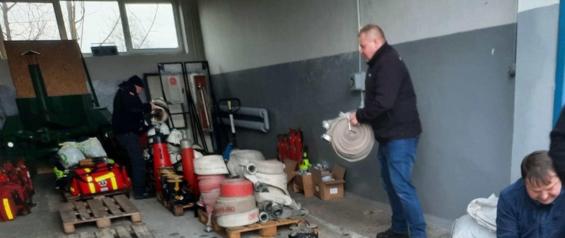 Garaż samochodowy, wewnątrz palety drewniane na których gromadzony jest sprzęt pożarniczy, na pierwszym palnie kartony w butami strażackimi oraz dwóch mężczyzn zabezpieczających węże do transportu
