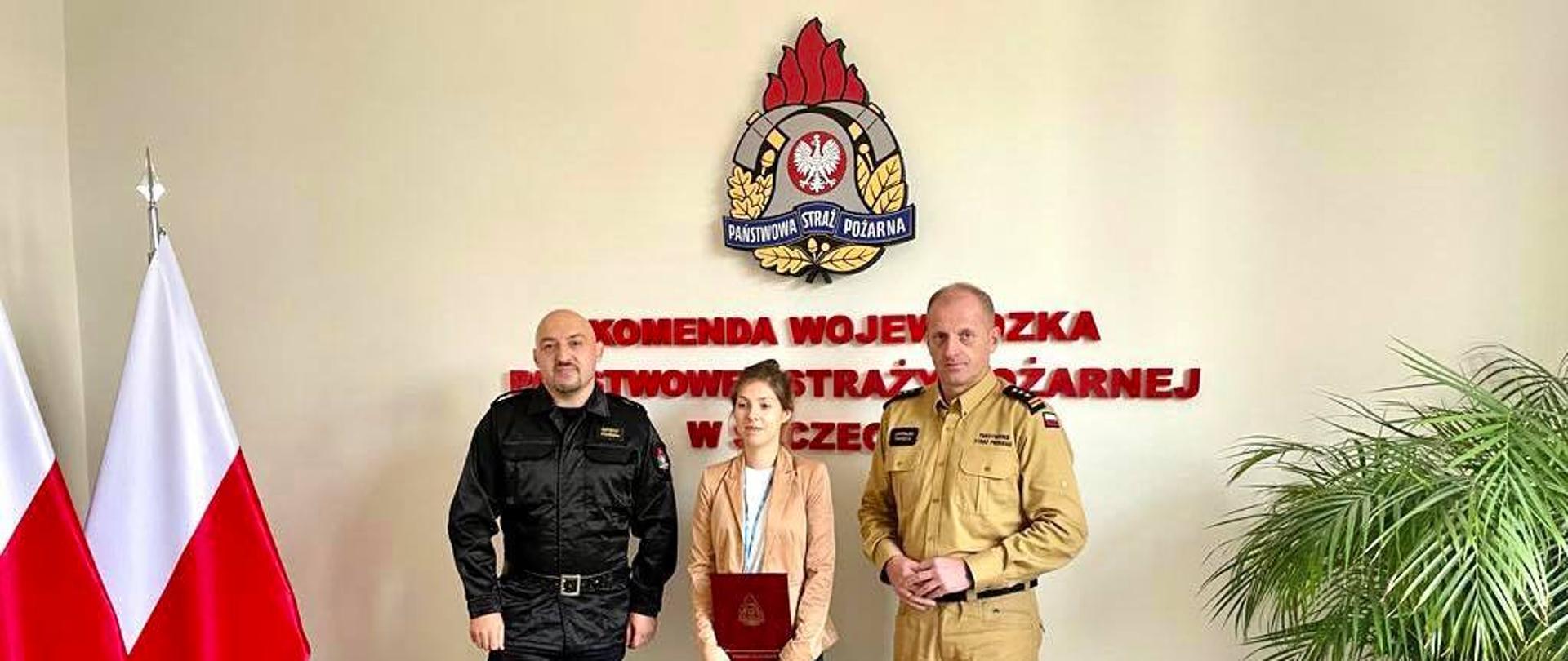 Uroczyste pożegnanie st. spec. Agaty Wronki. Na zdjęciu na tle ściany z logiem PSP widoczni są od lewej Naczelnik Wydziału Kadr mł. kpt. mgr Krzysztof Stelmasiak, st. spec. Agata Wronka oraz Zachodniopomorski Komendant Wojewódzki Państwowej Straży Pożarnej st. bryg. mgr inż. Jarosław Tomczyk.