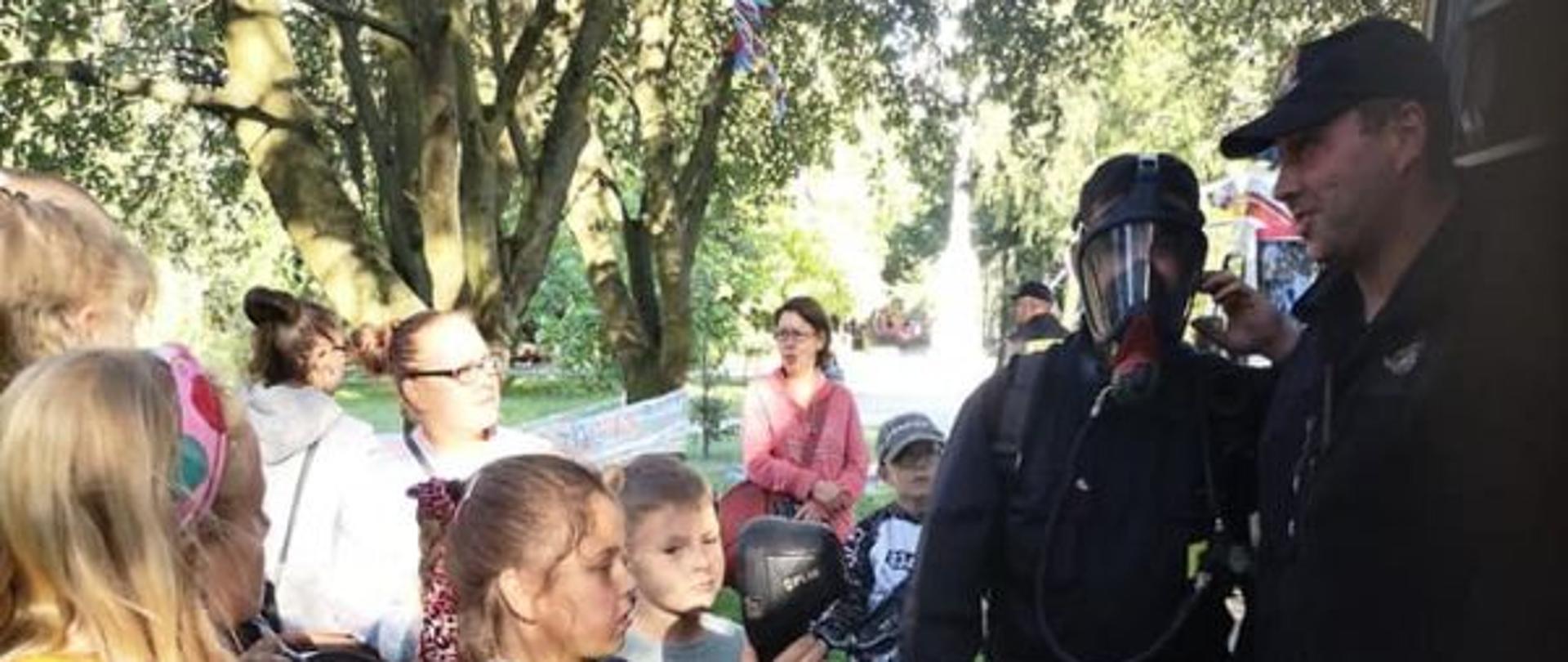 Zdjęcie grupowe. Strażacy stoją przy wozie z boku.Przed strażakami grupka dzieci.Zdjęcie wykonane w parku.Dzieci stoją i słuchają strażaków.Jeden z nich ma na sobie maskę aparatu ODO.
