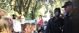 Zdjęcie grupowe. Strażacy stoją przy wozie z boku. Przed strażakami grupka dzieci.Zdjęcie wykonane w parku. Dzieci stoją i słuchają strażaków. Jeden z nich ma na sobie maskę aparatu ODO.