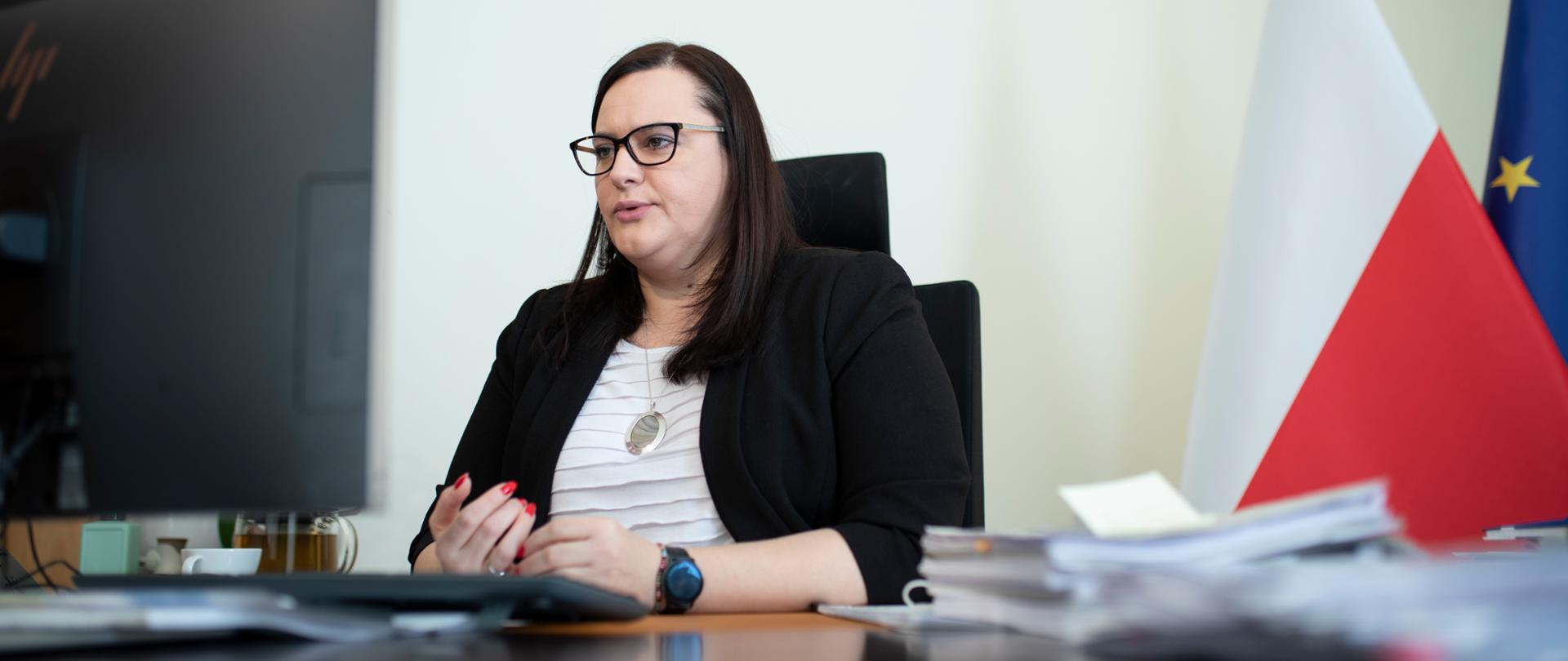 Wiceminister Małgorzata Jarosińska-Jedynak siedzi przy biurku przed monitorem komputera. Obok niej dwie flagi PL i UE.