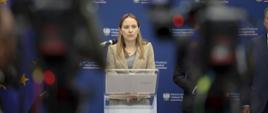 Minister Katarzyna Pełczyńska-Nałęcz stoi przy pulpicie z mikrofonem, za jej plecami ścianka z logo MFiPR