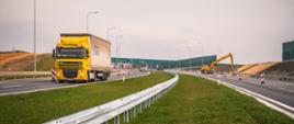 Ciężarówka jedzie nowo wybudowanym odcinkiem drogi ekspresowej w trakcie prac wykończeniowych; po bokach widoczne bariery energochłonne, oznakowanie robót drogowych oraz koparka na sąsiedniej jezdni.
