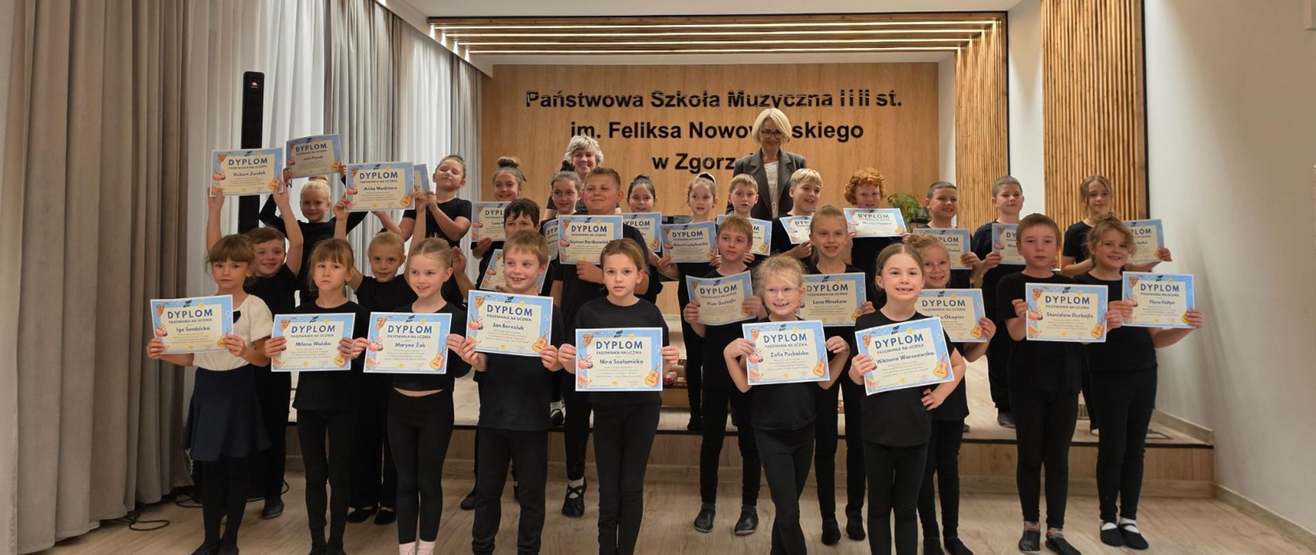 Grupa pierwszoklasistów w czarnych strojach rytmicznych stoi przy scenie auli koncertowej i trzyma w rękach pamiątkowe dyplomy pasowania na ucznia. Za dziećmi stoi nauczyciel rytmiki oraz dyrektor szkoły. Na ścianie widnieje napis z nazwą Państwowej Szkoły Muzycznej I i II st. im. Feliksa Nowowiejskiego w Zgorzelcu.