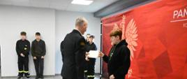 Strażak w mundurze wyjściowym gratuluje chłopakowi i wręcza dyplom. W prawej części zdjęcia czerwony baner PSP. W tle przed białą ścianą stoją inni uczestnicy turnieju. 