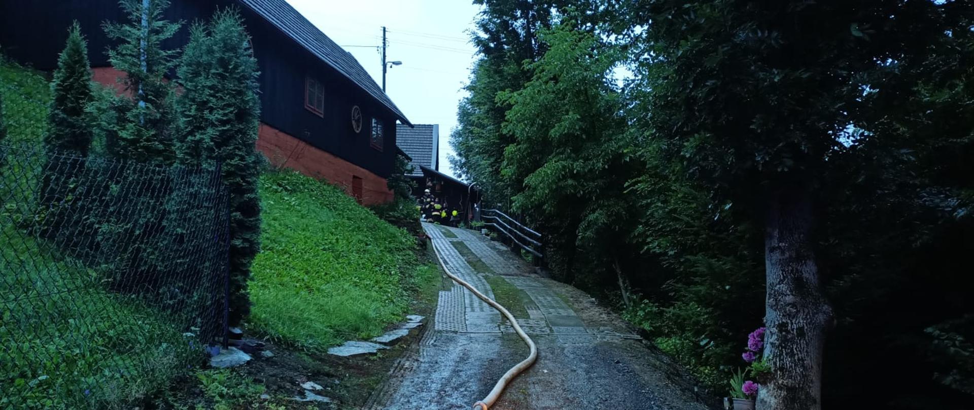 Linia wężowa prowadzona w kierunku zabudowań. W tle strażacy.