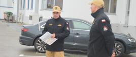 Ćwiczenia w GOBARTO. Zdjęcie przedstawia plac na terenie firmy. Na tle budynku i samochodu stoi dwóch strażaków PSP. Ubrani sa w umundurowanie służbowe w kolorze piaskowym oraz w czarne polary Dokonują podsumowania ćwiczeń.