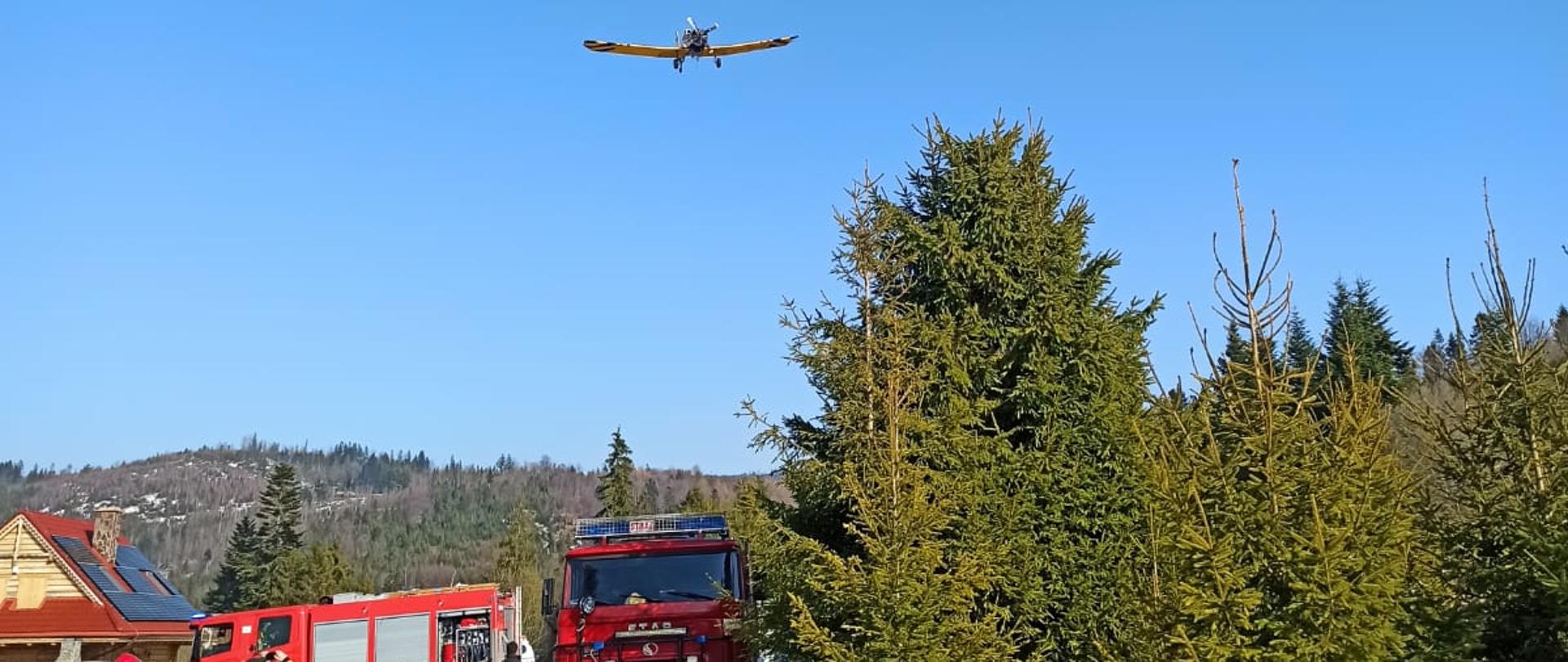 Samochody strażackie a nad nimi nisko przelatuje samolot Dromader przystosowany do gaszenia pożarów lasów