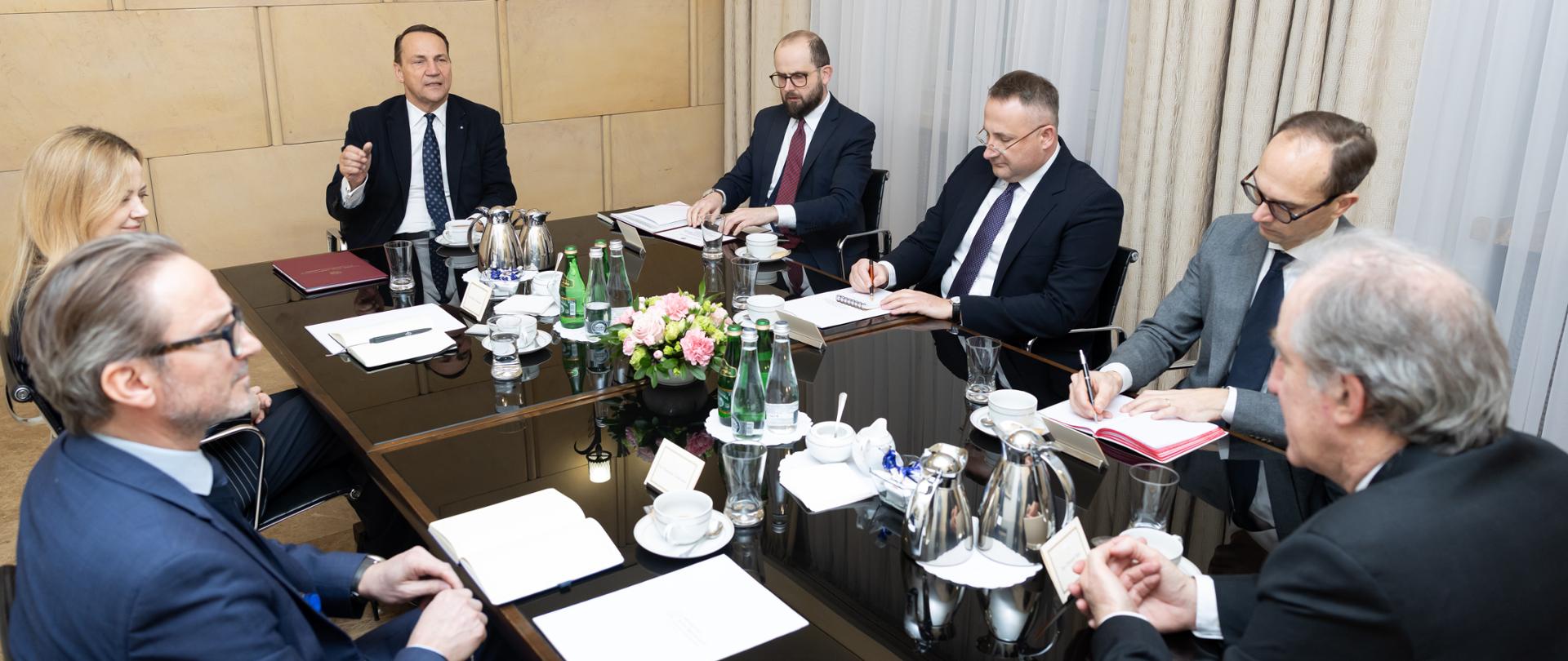 6 mężczyzn i jedna kobieta w formalnych strojach siedzących przy stole i rozmawiających.