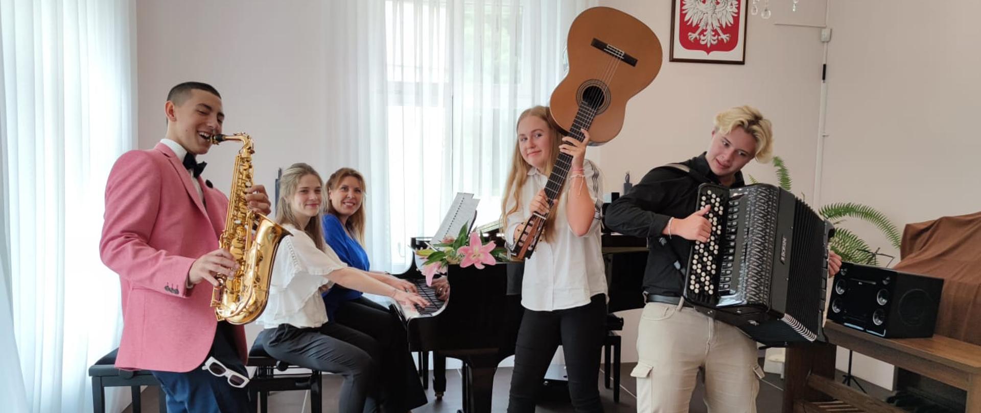 Zdjęcie przedstawia grupę uczniów saksofonista, gitarzystka i akordeonista wraz z nauczycielkami grającymi na fortepianie. 