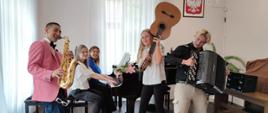 Zdjęcie przedstawia grupę uczniów saksofonista, gitarzystka i akordeonista wraz z nauczycielkami grającymi na fortepianie. 