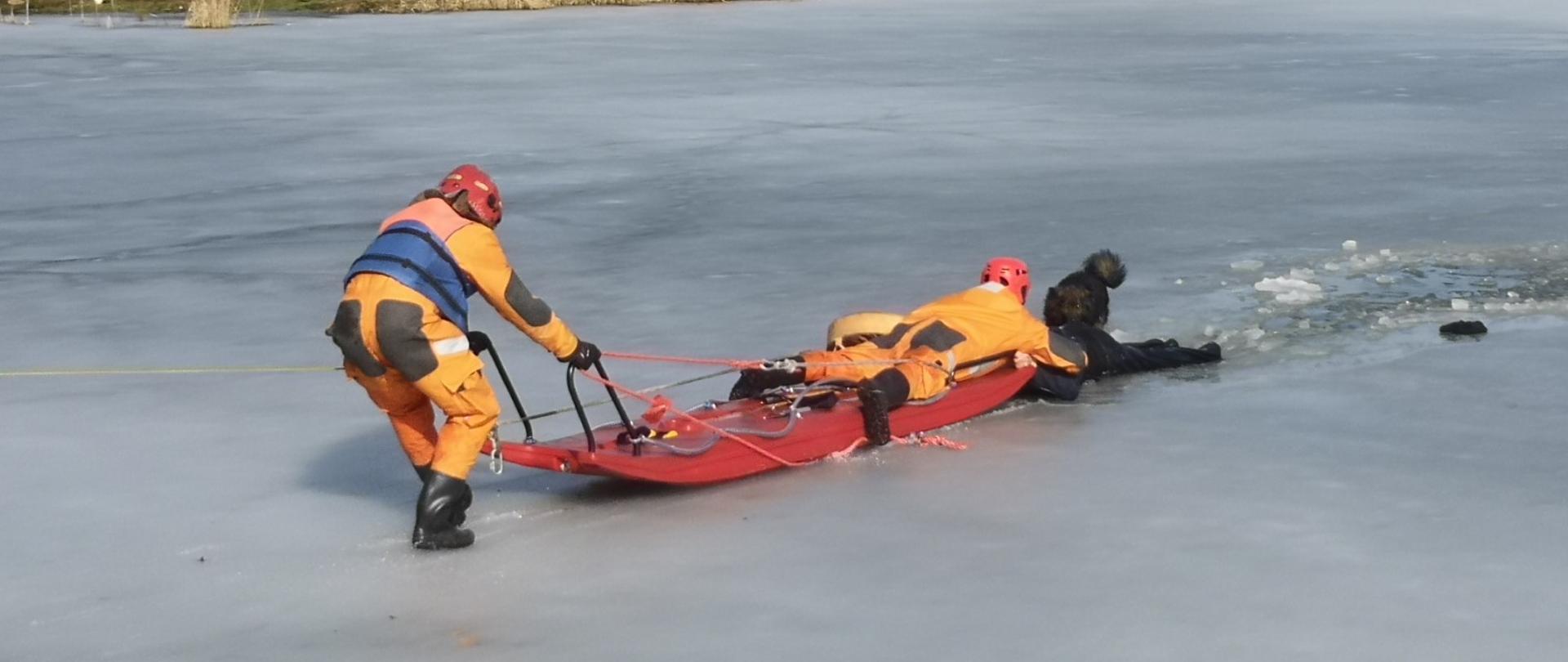 Zdjęcie przedstawia dwóch ratowników PSP z saniami wodno-lodowymi w trakcie ewakuacji mężczyzny z wody.