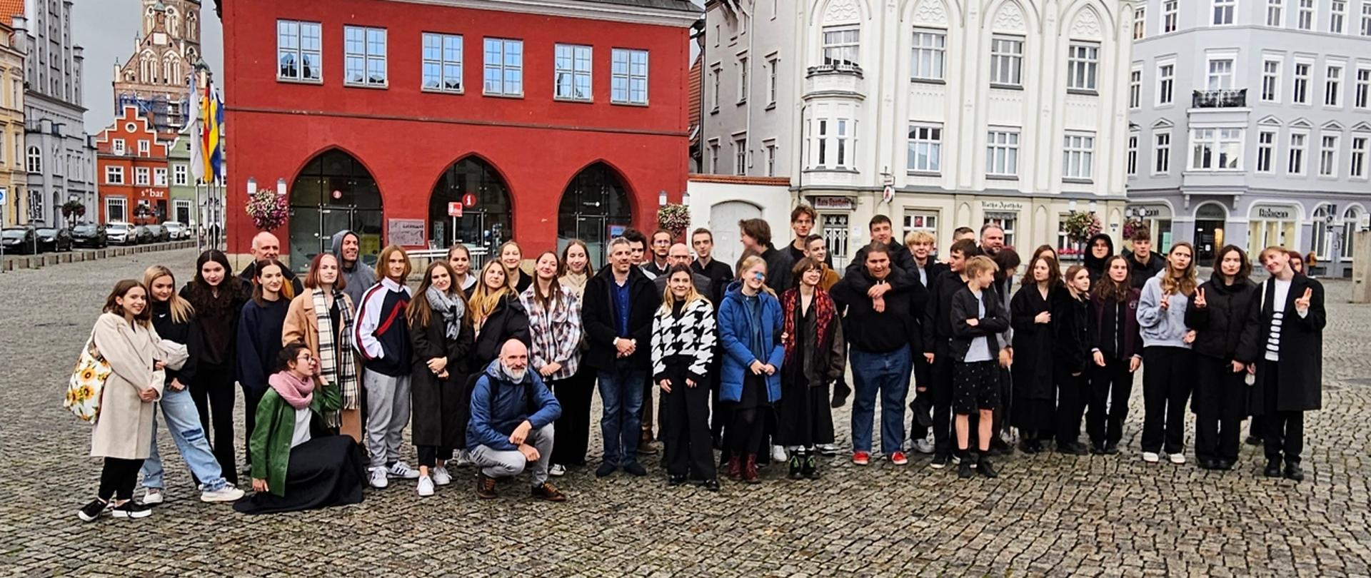 zdjęcie przedstawia uczestników warsztatów na środku rynku zabytkowej miejscowości, w tle kamienice i ratusz