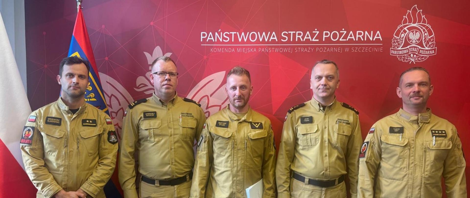 Pięciu funkcjonariuszy Państwowej Straży Pożarnej w beżowych mundurach służbowych stoi w szeregu na tle czerwonej ścianki z logotypem Komendy Miejskiej PSP w Szczecinie. Po lewej stronie widoczne są flagi Polski i Państwowej Straży Pożarnej.