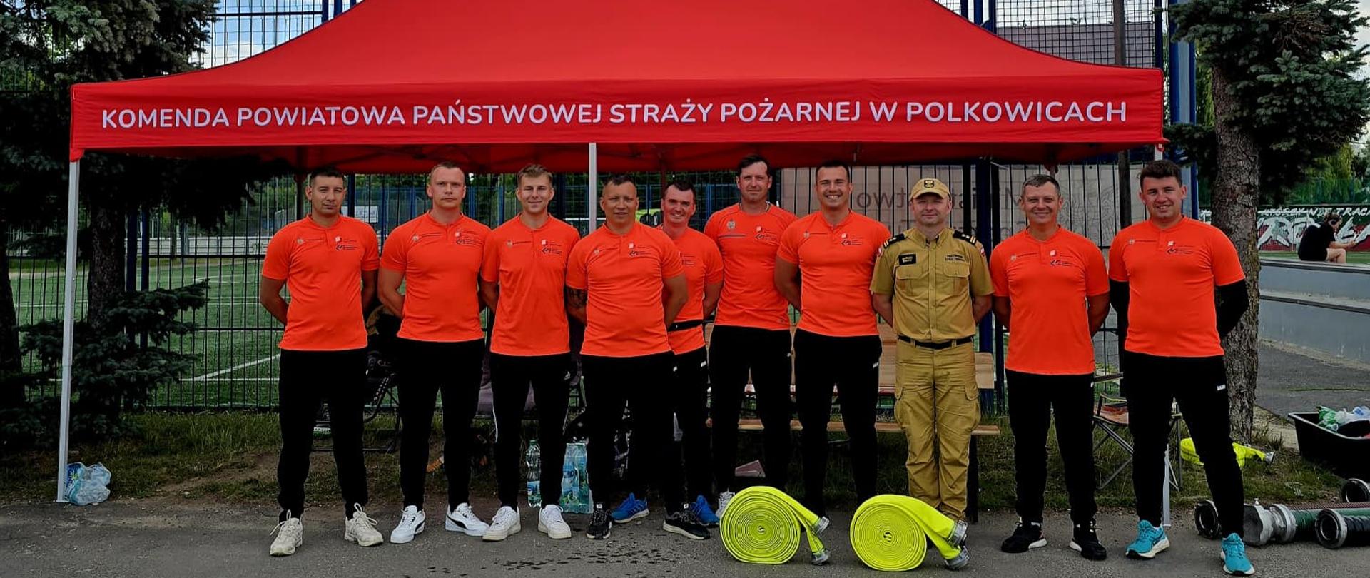 Na zdjęciu przedstawiona jest grupa mężczyzn – strażaków oraz reprezentantów – pozujących do wspólnego zdjęcia na świeżym powietrzu, prawdopodobnie podczas zawodów sportowo-pożarniczych lub wydarzenia plenerowego.
---
## Opis postaci
* **Ubiór sportowy:** Większość mężczyzn (dziewięciu z dziesięciu) ubrana jest w jednolite stroje sportowe składające się z jaskrawopomarańczowych koszulek z krótkim rękawem (z logo na piersi) oraz długich, czarnych spodni sportowych i butów sportowych.
* **Ubiór służbowy:** Jedna osoba (czwarta od prawej) wyróżnia się ubiorem – ma na sobie piaskowy (beżowy) mundurowy strój służbowy Państwowej Straży Pożarnej, czapkę z daszkiem w tym samym kolorze oraz czarny pas.
---
## Otoczenie i rekwizyty
* **Namiot:** Mężczyźni stoją w jednym rzędzie pod dużym, czerwonym namiotem rozstawowym. Na przedniej falbanie namiotu widnieje duży, biały napis w języku polskim: **„KOMENDA POWIATOWA PAŃSTWOWEJ STRAŻY POŻARNEJ W POLKOWICACH”**.
* **Sprzęt strażacki:** Na ziemi, przed stojącymi mężczyznami, leżą dwie zwinięte w kręgi, jaskrawożółte (limonkowe) linie węży strażackich. Po prawej stronie na ziemi widoczne są również metalowe rozdzielacze lub nasady.
* **Tło:** Za namiotem znajduje się wysokie ogrodzenie z siatki (prawdopodobnie otaczające boisko sportowe), a po bokach rosną wysokie drzewa iglaste (świerki). Pogoda na zdjęciu wydaje się pochmurna.