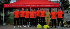 Na zdjęciu przedstawiona jest grupa mężczyzn – strażaków oraz reprezentantów – pozujących do wspólnego zdjęcia na świeżym powietrzu, prawdopodobnie podczas zawodów sportowo-pożarniczych lub wydarzenia plenerowego.
---
## Opis postaci
* **Ubiór sportowy:** Większość mężczyzn (dziewięciu z dziesięciu) ubrana jest w jednolite stroje sportowe składające się z jaskrawopomarańczowych koszulek z krótkim rękawem (z logo na piersi) oraz długich, czarnych spodni sportowych i butów sportowych.
* **Ubiór służbowy:** Jedna osoba (czwarta od prawej) wyróżnia się ubiorem – ma na sobie piaskowy (beżowy) mundurowy strój służbowy Państwowej Straży Pożarnej, czapkę z daszkiem w tym samym kolorze oraz czarny pas.
---
## Otoczenie i rekwizyty
* **Namiot:** Mężczyźni stoją w jednym rzędzie pod dużym, czerwonym namiotem rozstawowym. Na przedniej falbanie namiotu widnieje duży, biały napis w języku polskim: **„KOMENDA POWIATOWA PAŃSTWOWEJ STRAŻY POŻARNEJ W POLKOWICACH”**.
* **Sprzęt strażacki:** Na ziemi, przed stojącymi mężczyznami, leżą dwie zwinięte w kręgi, jaskrawożółte (limonkowe) linie węży strażackich. Po prawej stronie na ziemi widoczne są również metalowe rozdzielacze lub nasady.
* **Tło:** Za namiotem znajduje się wysokie ogrodzenie z siatki (prawdopodobnie otaczające boisko sportowe), a po bokach rosną wysokie drzewa iglaste (świerki). Pogoda na zdjęciu wydaje się pochmurna.