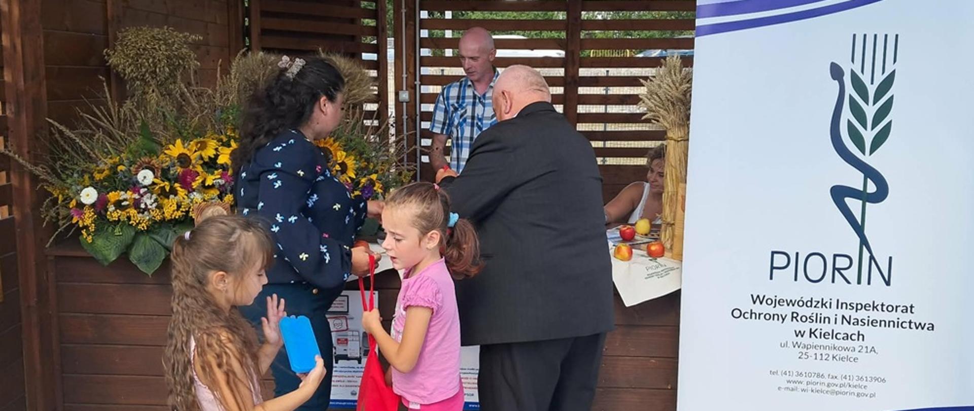 W centralnej części zdjęcia są widoczne osoby zainteresowane stoiskiem, mężczyzna oraz kobieta. Mężczyzna ubrany w ciemny garnitur kobieta ubrana w koszulę o ciemnym odcieniu. Na pierwszym planie widoczne jest dwoje dzieci, dziewczynki ubrane w kolorach białym oraz różowym. Na dalszym planie widoczni są pracownicy inspekcji mężczyzna i kobieta udzielający porad zainteresowanym. Po prawej stronie zdjęcia widoczny jest roll-up Państwowej Inspekcji ochrony Roślin i Nasiennictwa w Kielcach, zielony kłos opleciony czarnym wężem na biało niebieskim tle. 