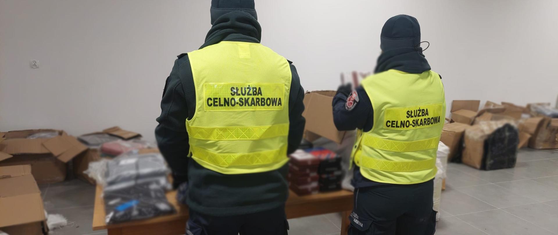 Dwoje funkcjonariuszy Służby Celno‑Skarbowej w żółtych kamizelkach odblaskowych stoi w magazynie wśród kartonów i zabezpieczonych towarów.