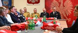 Za stołem siedzą zaproszeni goście, na stole znajdują się torby z logiem PSP z tyłu godło Polski oraz sztandar PSP