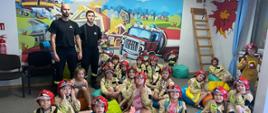 Grupa dzieci biorących udział w półkoloniach na sali edukacyjnej OGNIK. Za nimi dwóch strażaków na tle grafiki strażackiej