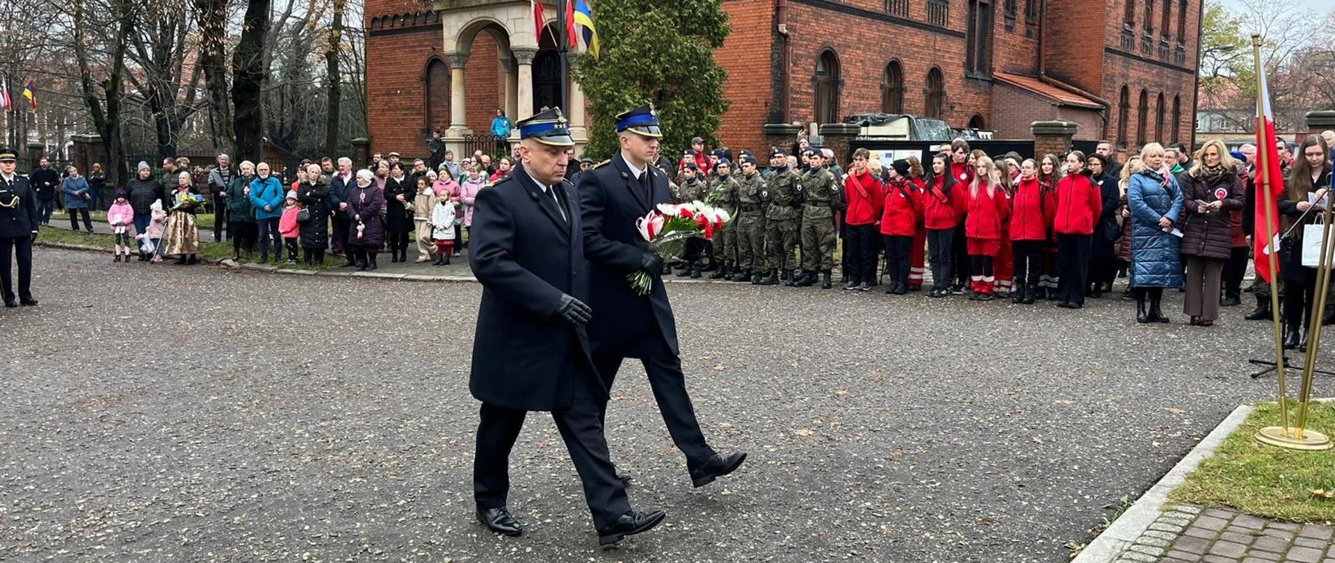 Na zdjęciu widocznych jest dwóch strażaków w umudurowaniu galowym, w płaszczach. Strażacy podczas marszu w kierunku tablicy „Pamięci tych, co grobów nie mają”. Jeden ze strażaków trzyma w rękach kwiaty w kolorze białym i czerwonym. W tle budynek kościoła, a przed nim zebrani uczestnicy uroczystości.