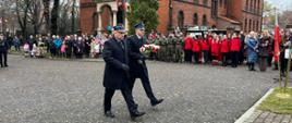 Na zdjęciu widocznych jest dwóch strażaków w umudurowaniu galowym, w płaszczach. Strażacy podczas marszu w kierunku tablicy „Pamięci tych, co grobów nie mają”. Jeden ze strażaków trzyma w rękach kwiaty w kolorze białym i czerwonym. W tle budynek kościoła, a przed nim zebrani uczestnicy uroczystości.
