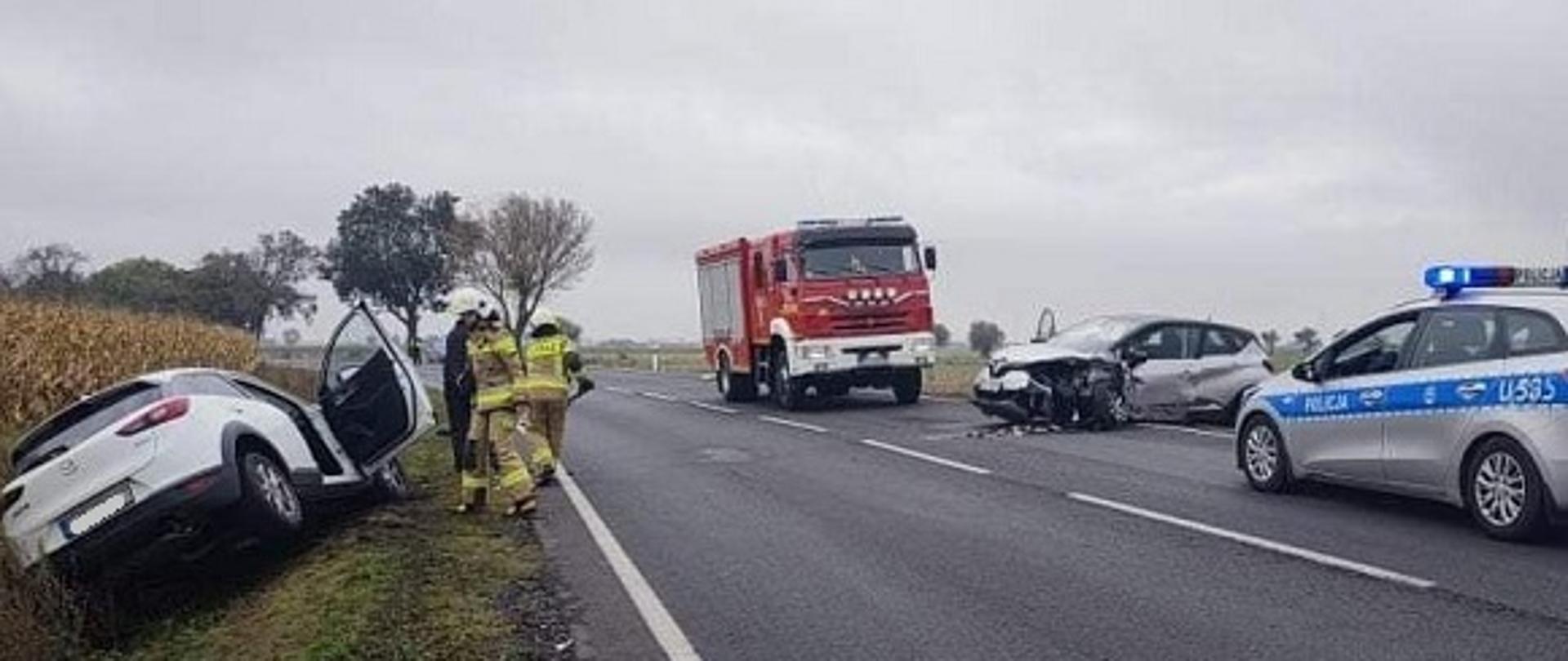 Działania na miejscu wypadku dwóch samochodów. na jednym pasie drogi krajowej pojazd straży pożarnej, policji oraz uszkodzony środek transportu. Drugi z pojazdów uczestniczących w zdarzeniu leży w rowie. Tuż obok pojazdów stoją strażacy realizujący działania. Droga zablokowana. 