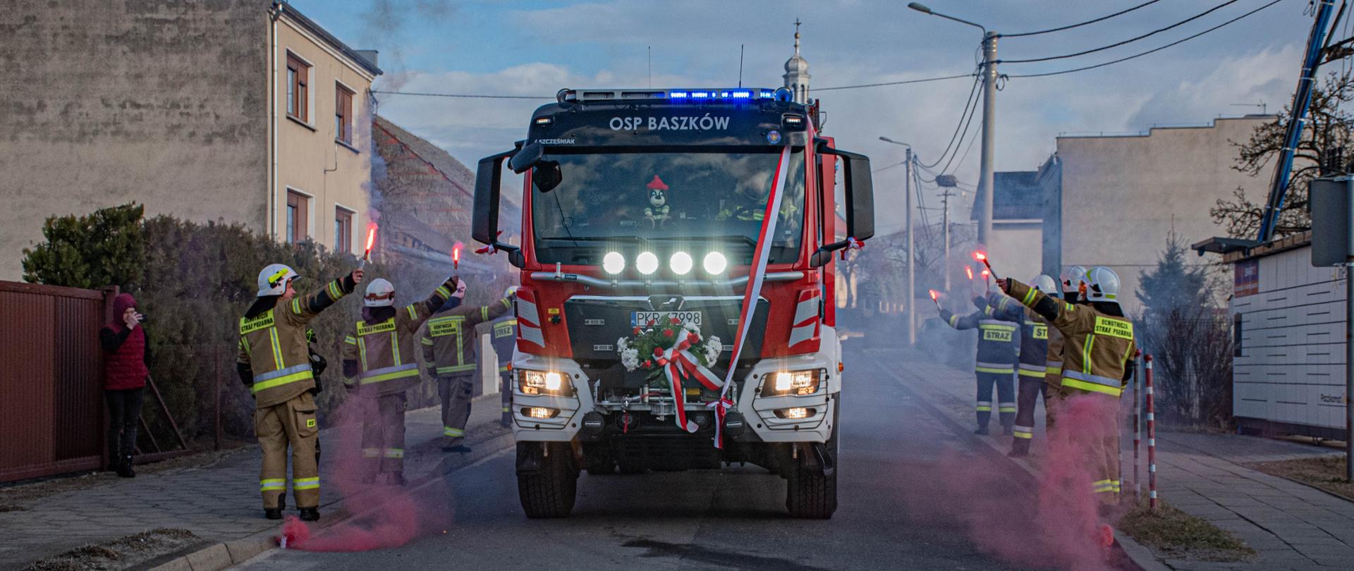 Na zdjęciu nowy pojazd jednostki OSP Baszków, który wjeżdża na teren miejscowości. Trasę dojazdu obstawiają druhowi jednostki w ubraniach bojowych trzymając w rękach race dymne podkreślając radość z doposażenia jednostki
