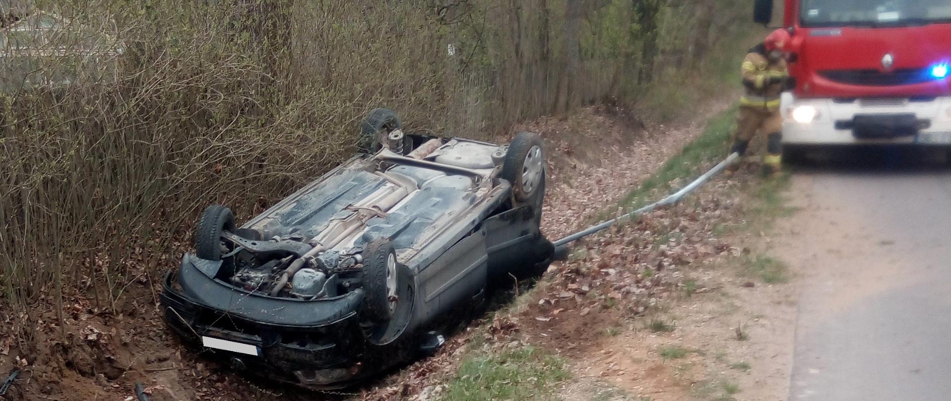 Samochód osobowy leżący na dachu w przydrożnym rowie. W tle strażak oraz wóz strażacki.