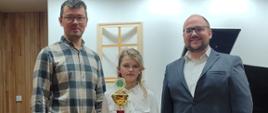 Zdjęcie przedstawia osoby stojące, trzymającą puchar uczennicę Zuzannę Wojciechowski pośrodku, po lewej akompaniator, pan Daniel Rybak, po prawej nauczyciel saksofonu, pan Adrian Masseli. 