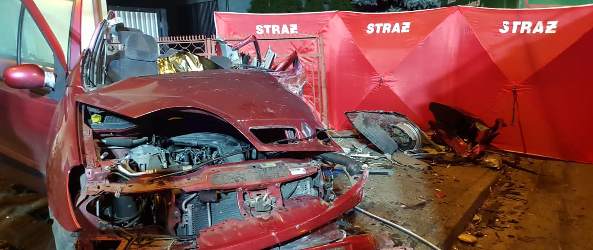 Czerwony samochód osobowy marki Reanult wjechał w płot posesji. Jest cały zgnieciony po wypadku. Za nim rozłożony parawan strażacki.
