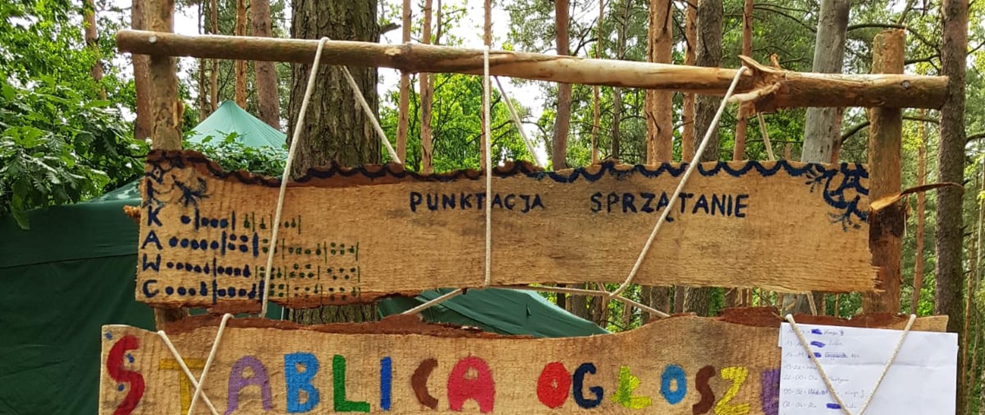 Zdjęcie przedstawia tablicę ogłoszeń w obozie harcerskim. 