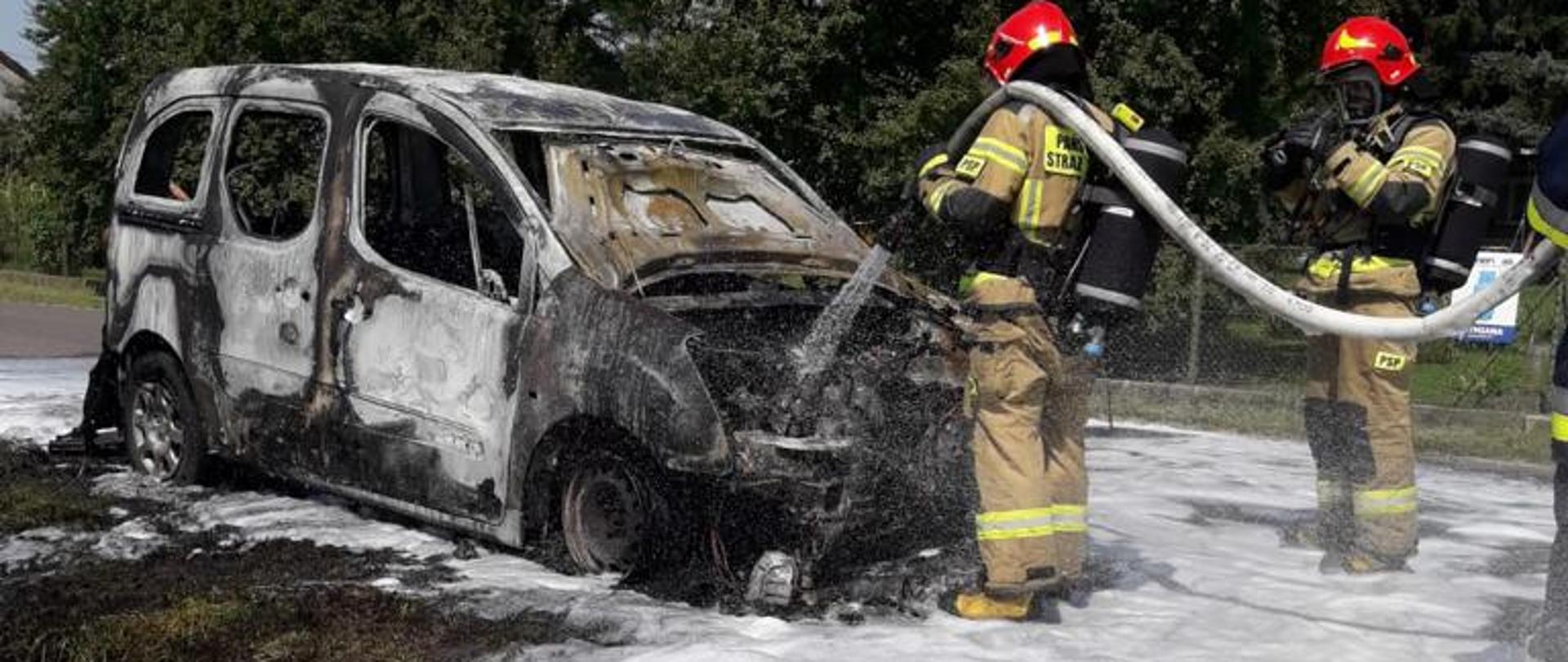 Na zdjęciu widoczni strażacy podające prąd wody, celem dogaszenia samochodu osobowego, samochód całkowicie spalony, widoczna piana gaśnicza.