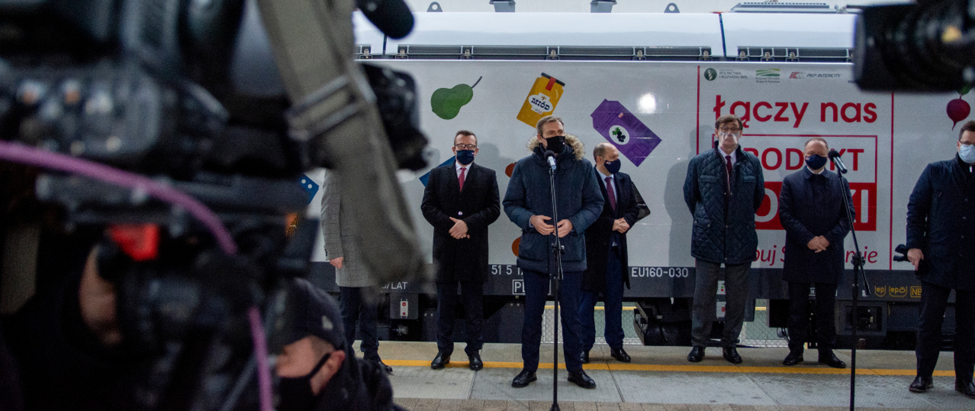 Briefing prasowy - przy mikrofonie wiceminister Maciej Małecki. Za nim stoją pozostali uczestnicy. W tle oklejona okolicznościowo lokomotywa