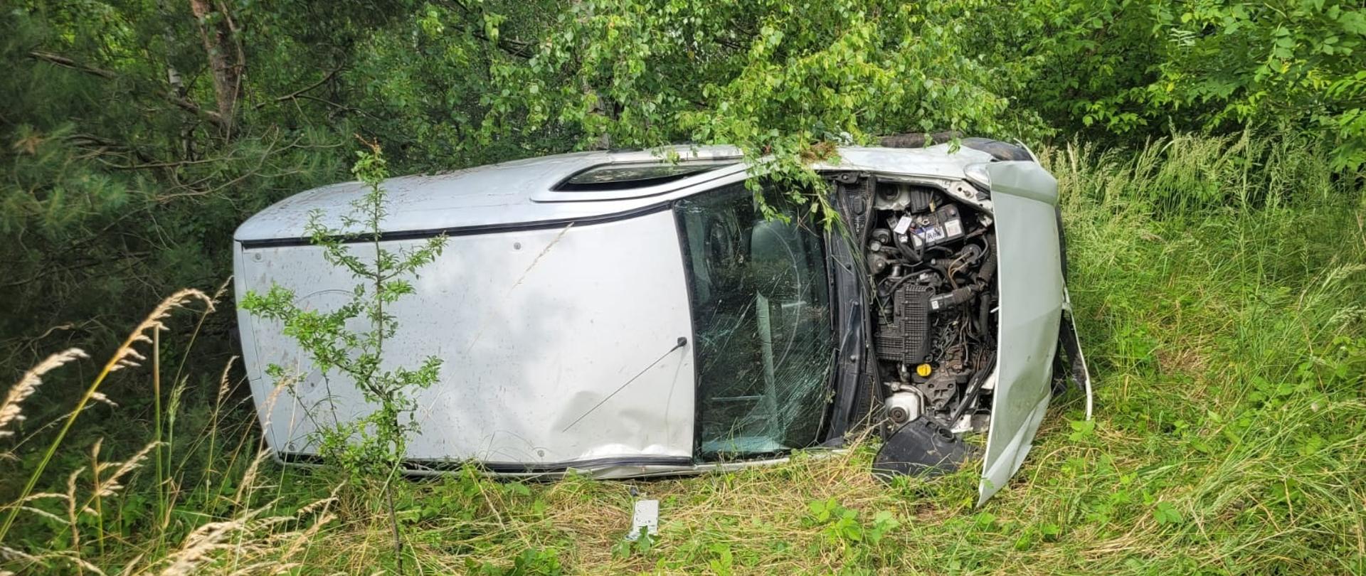 Na zdjęciu widoczny jest srebrny samochód osobowy, który uległ wypadkowi drogowemu i leży na boku w zaroślach, poza jezdnią, w otoczeniu gęstej roślinności i drzew. Pojazd jest częściowo uszkodzony – widoczna jest zniszczona przednia część, z otwartą maską ukazującą silnik. Szyby są popękane, a wokół samochodu znajdują się ślady przejazdu przez trawę i roślinność. Scena wskazuje na możliwą utratę panowania nad pojazdem i zjazd z drogi do lasu lub zalesionego pobocza.
