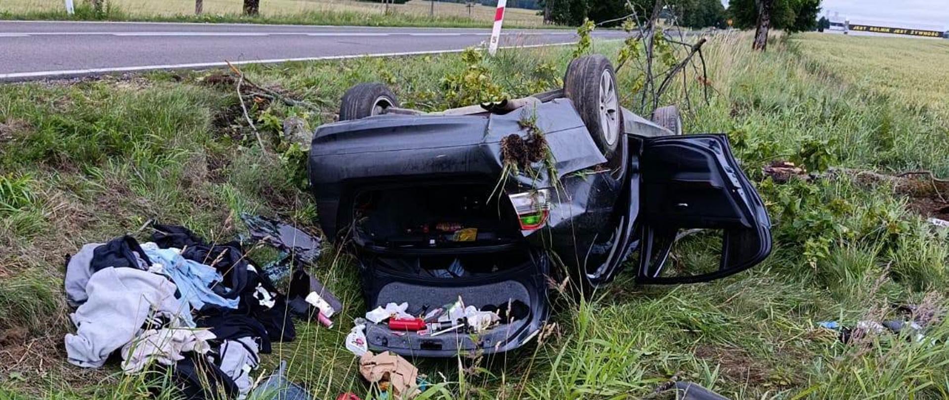 Samochód osobowy na dachu na poboczu drogi. Otwarte drzwi i bagażnik. Porozrzucane elementy wyposażenia auta.