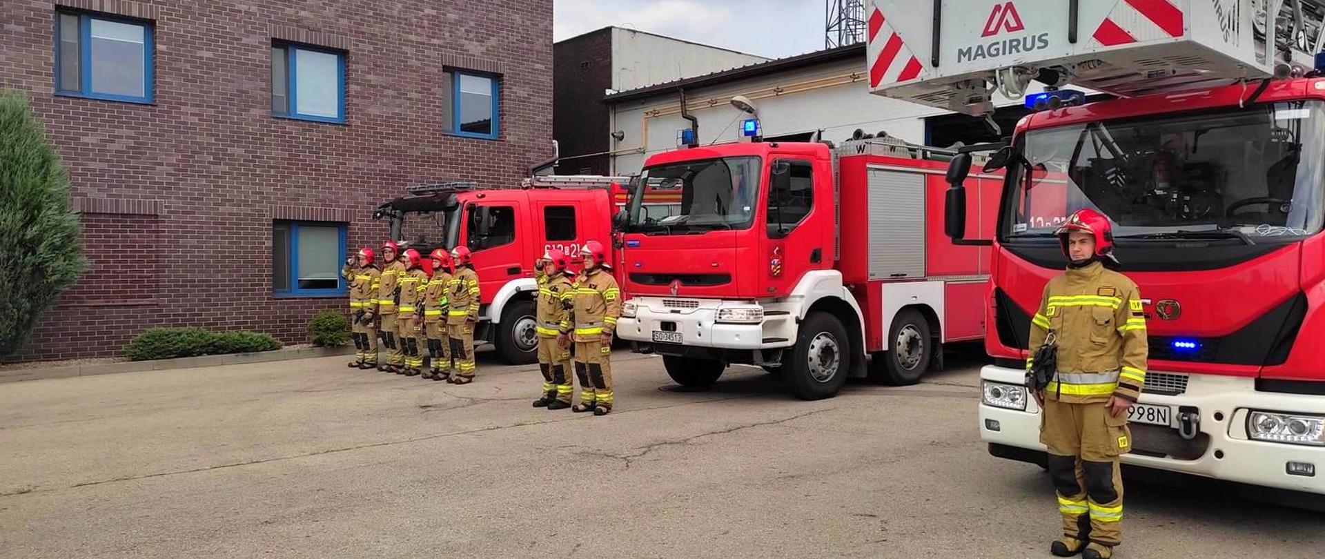 Na zdjęciu zmiana służbowa w umundurowaniu specjalnym na tle samochodów pożarniczych z włączonymi sygnałami świetlnymi 