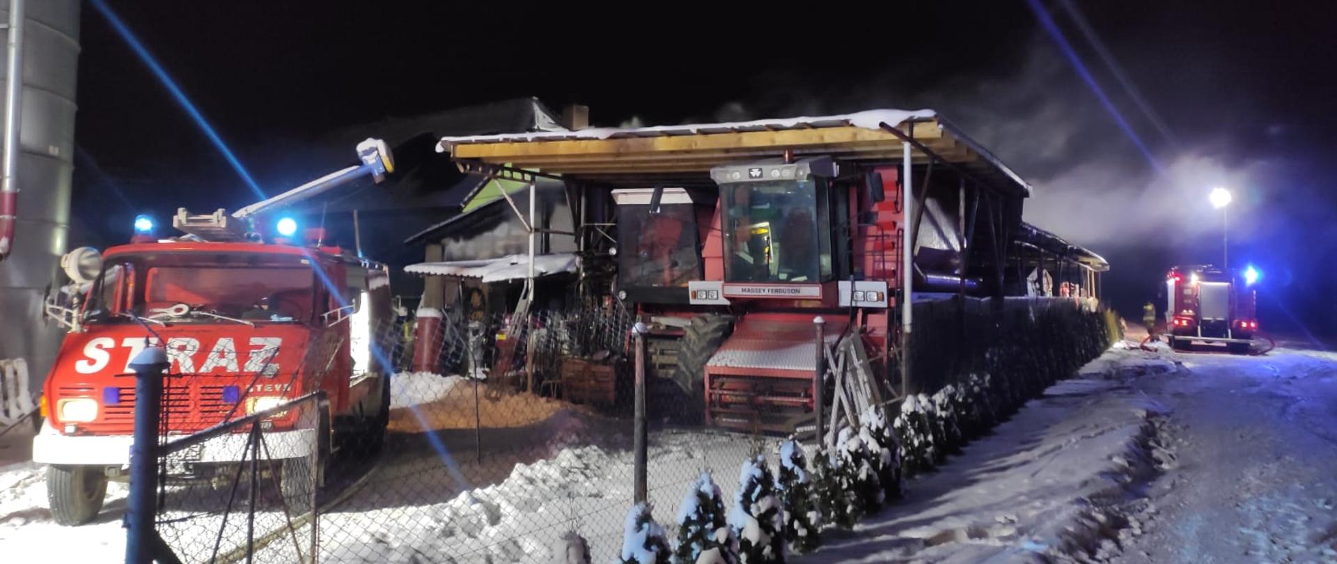 Obraz przedstawia dwa pojazdy pożarnicze z włączonymi światłami alarmowymi. Pora nocna. W środkowej części wiata pod którą stoją dwa kombajny. Nad wiatą widoczny unoszący się dym.